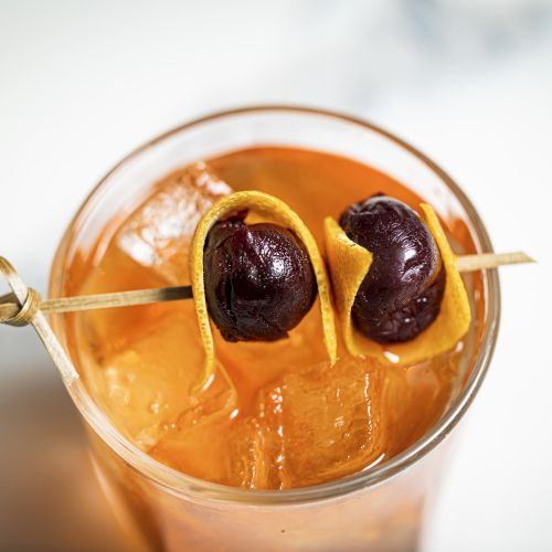 A glass filled with an orange-coloured drink and ice, garnished with two cherries and a citrus peel on a cocktail pick in this image.