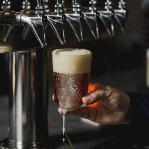 A person is pouring a glass of beer from a row of taps, with the beer forming a foam head at the top.