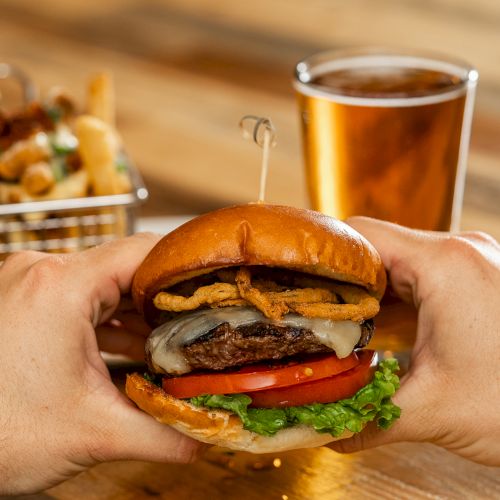 A person is holding a burger with lettuce, tomato, cheese, and crispy onions. In the background are fries and a glass of beer.