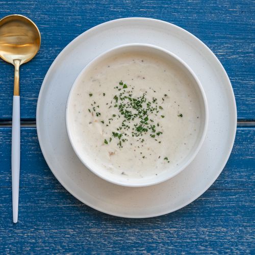 A bowl of creamy soup garnished with chopped herbs, served on a white plate with a gold spoon set on a blue wooden surface.