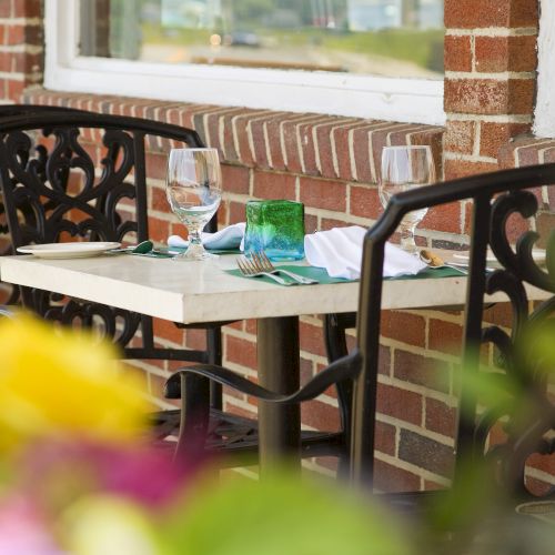 An outdoor table set for two with glassware, cutlery, and green napkins, next to a red brick wall, surrounded by blurred flowers in the foreground.