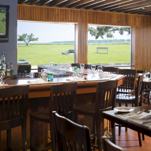 An upscale bar and dining area with wooden furniture, neatly set tables, a large TV displaying tennis, and large windows showing a scenic outdoor view.