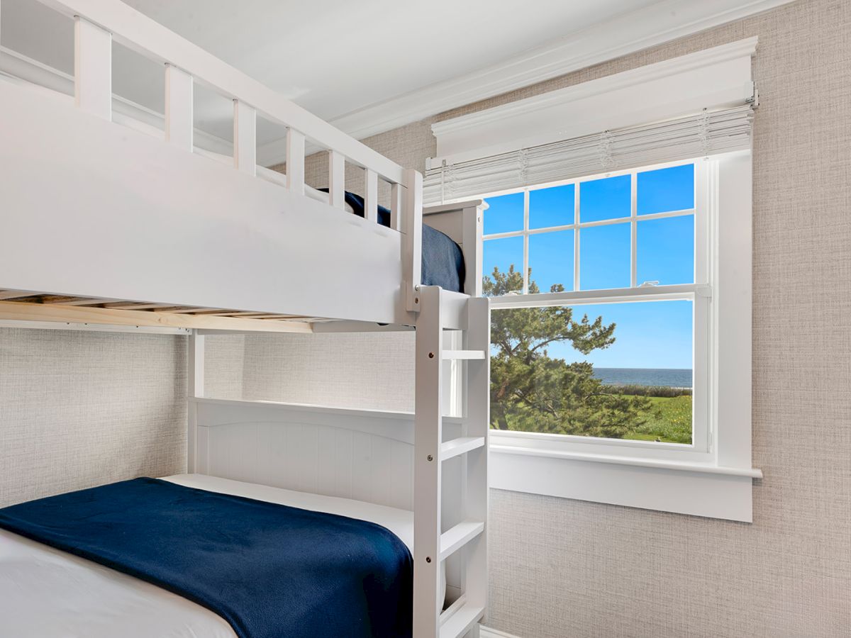 A bedroom with white bunk beds, blue bedding, a window with a view of greenery and the ocean under a bright blue sky.