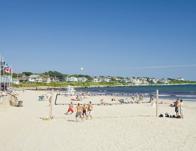 A beach scene with people playing volleyball, lounging, and walking. Flags are displayed on the left, and houses are visible in the background.