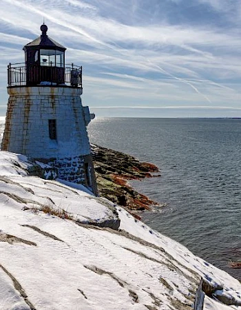 A lighthouse stands on a snowy cliff overlooking a calm sea under a sky with streaky clouds.