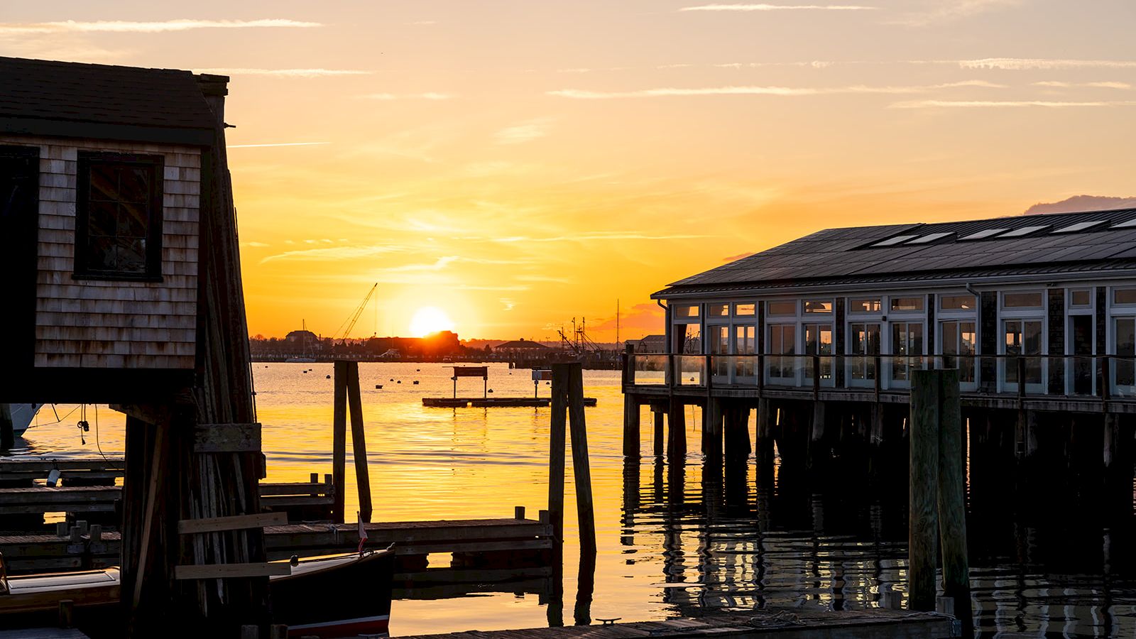 The image shows a sunset over a harbor with wooden buildings and docks, water reflecting the colorful sky.