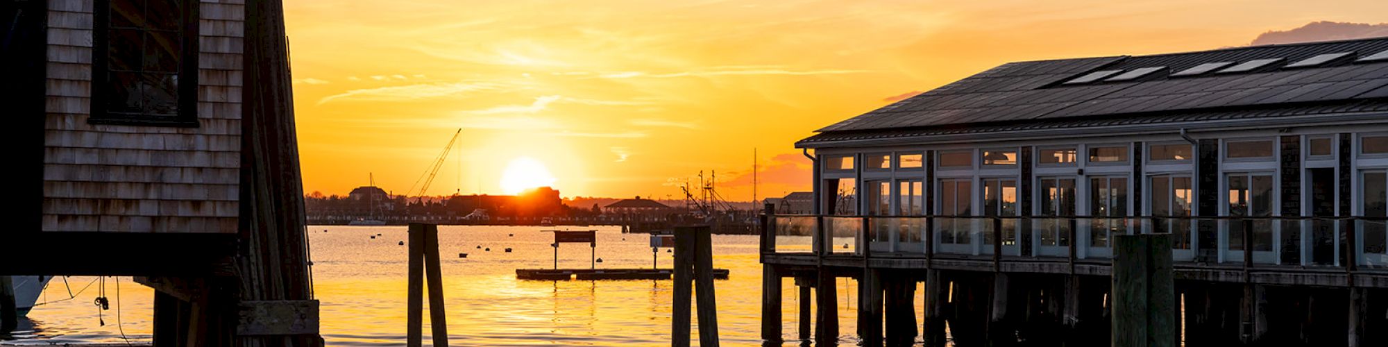 The image shows a sunset over a harbor with wooden buildings and docks, water reflecting the colorful sky.