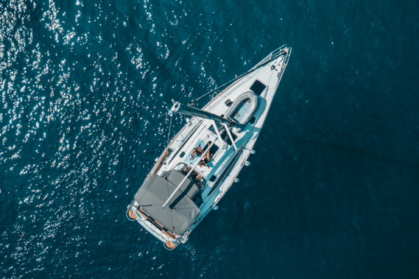Aerial view of a white sailboat with several people onboard, cruising on a clear blue sea. The boat has sunshades and appears to be in leisure mode.