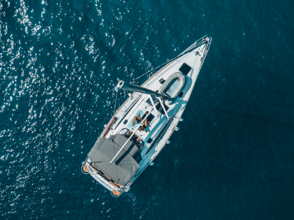 Aerial view of a white sailboat with several people onboard, cruising on a clear blue sea. The boat has sunshades and appears to be in leisure mode.