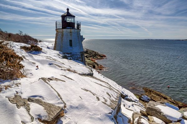 A lighthouse stands on a rocky, snow-covered shore with the sea stretching out beside it under a cloudy blue sky.