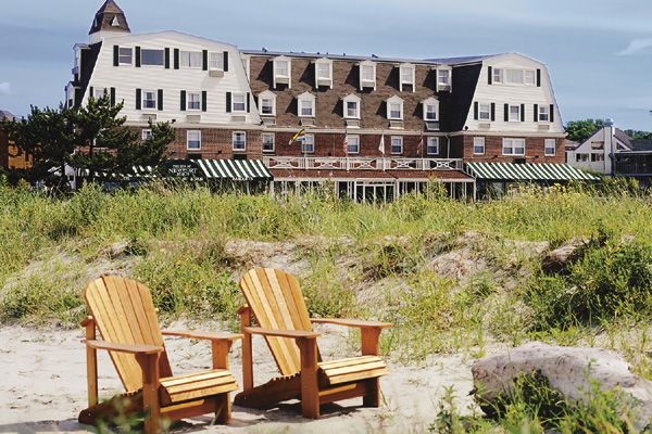 Two wooden chairs on a sandy beach with a grassy area in front of a large building under a blue sky.