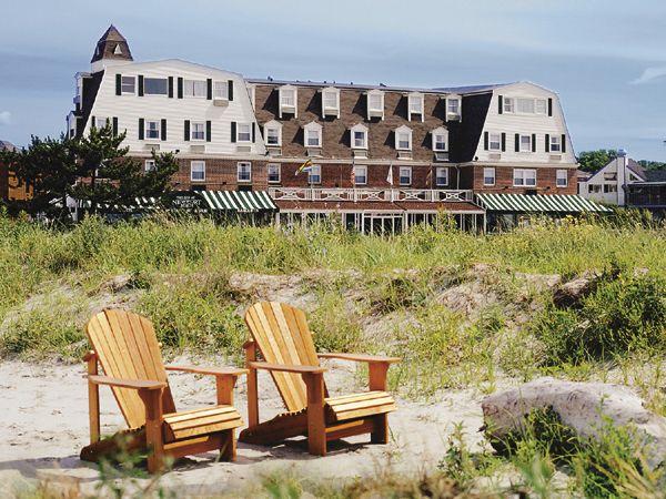Two wooden chairs on a sandy beach with a grassy area in front of a large building under a blue sky.