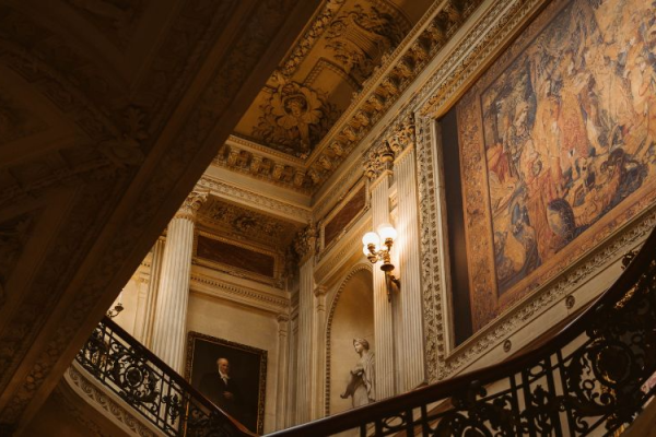 An ornate staircase with intricate railings, a large tapestry on the wall, decorative ceilings, and columns in a grand interior setting.