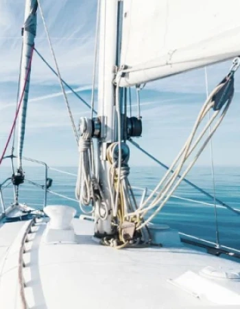 A view from the deck of a sailboat on calm waters, with visible ropes, mast, and sails under a clear blue sky.