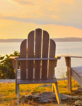 Two wooden chairs facing a scenic sunset over a body of water, surrounded by grass and shrubs, creating a peaceful atmosphere.