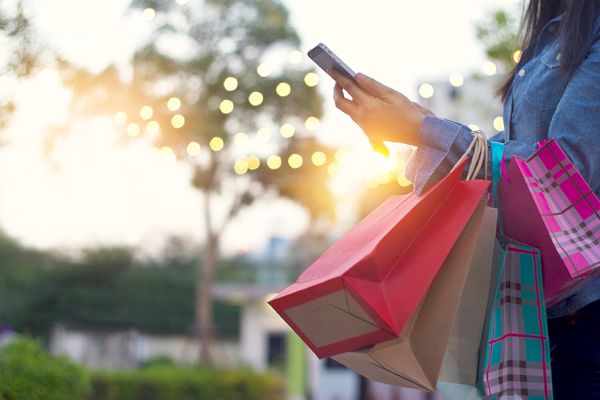 A person holding colorful shopping bags and a smartphone outdoors, with blurred lights and a sunset in the background.
