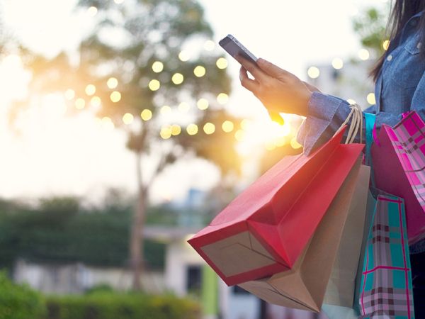 A person holding colorful shopping bags and a smartphone outdoors, with blurred lights and a sunset in the background.