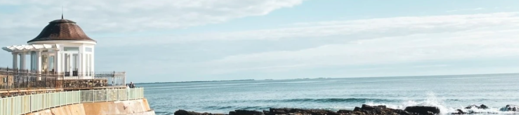 A coastal scene with a gazebo, rocky shore, and calm sea under a blue sky with clouds.