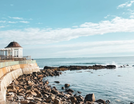A coastal scene with a gazebo, rocky shore, and calm sea under a blue sky with clouds.