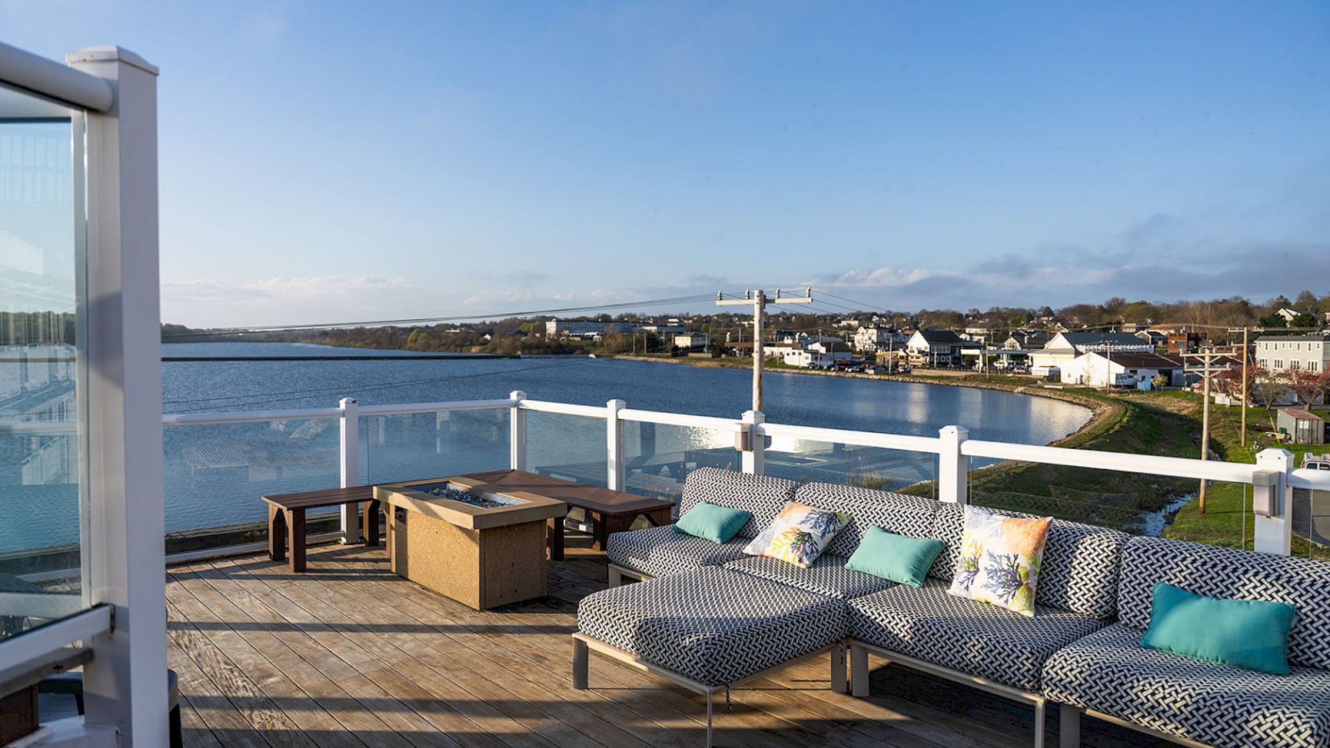 A sunny coastal deck with wicker sectional seating, colorful cushions, a hot tub, and a view of calm water and houses across the bay.