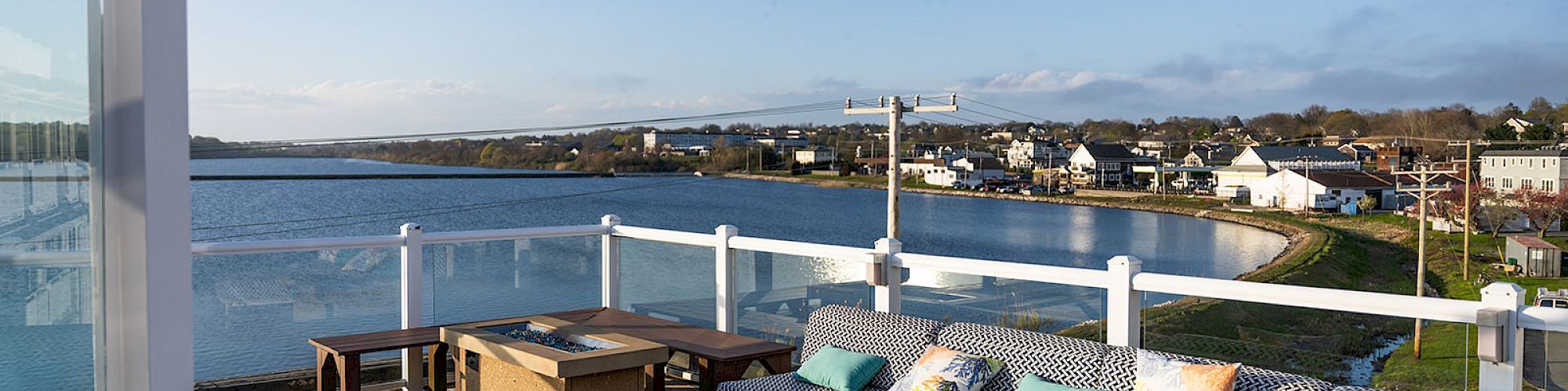 A sunny coastal deck with wicker sectional seating, colorful cushions, a hot tub, and a view of calm water and houses across the bay.