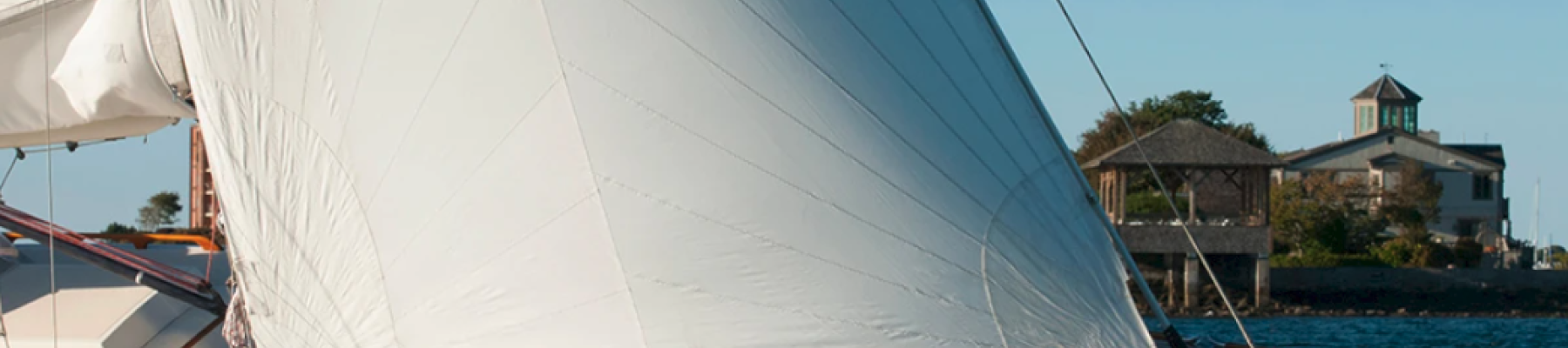 A white-sailed sailboat cuts through blue water near shore with a small building visible in the background.