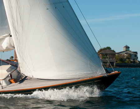 A white-sailed sailboat cuts through blue water near shore with a small building visible in the background.
