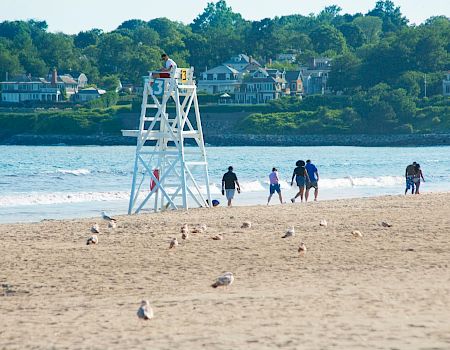 On a sunny beach, lifeguard tower stands as a group of people stroll along the shoreline near calm waves, with houses in the background.