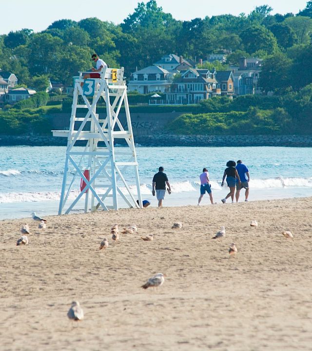 On a sunny beach, lifeguard tower stands as a group of people stroll along the shoreline near calm waves, with houses in the background.