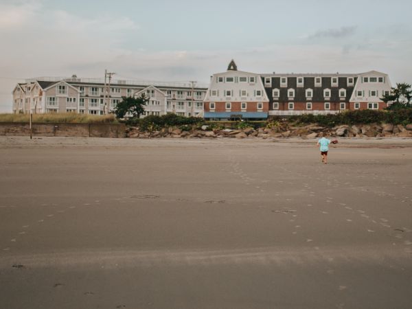 A lone person walks along a wide, empty beach with rows of pastel beach houses in the background under a calm sky.