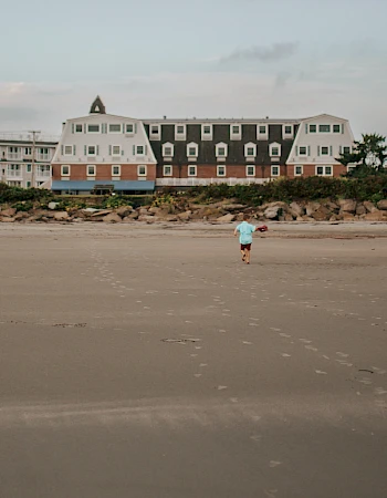A quiet beach with wide sand, a row of pastel seaside houses in the background, and a lone person walking near the shore.