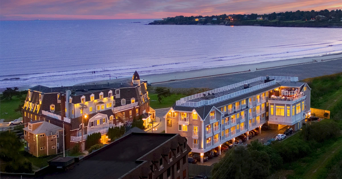 A seaside resort town with multi-story hotel buildings lit up at dusk, facing a calm beach and shoreline, overlooking the water and distant coastline.