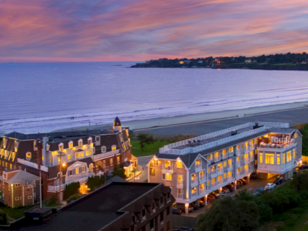 Coastal resort town at dusk with lit seaside buildings, calm waves, and a colorful sunset over the horizon.