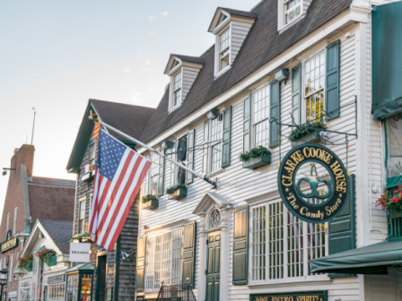 A charming street with white clapboard buildings, an American flag, and a round &ldquo;Clarke Cooke House&rdquo; candy shop sign, sunny day.
