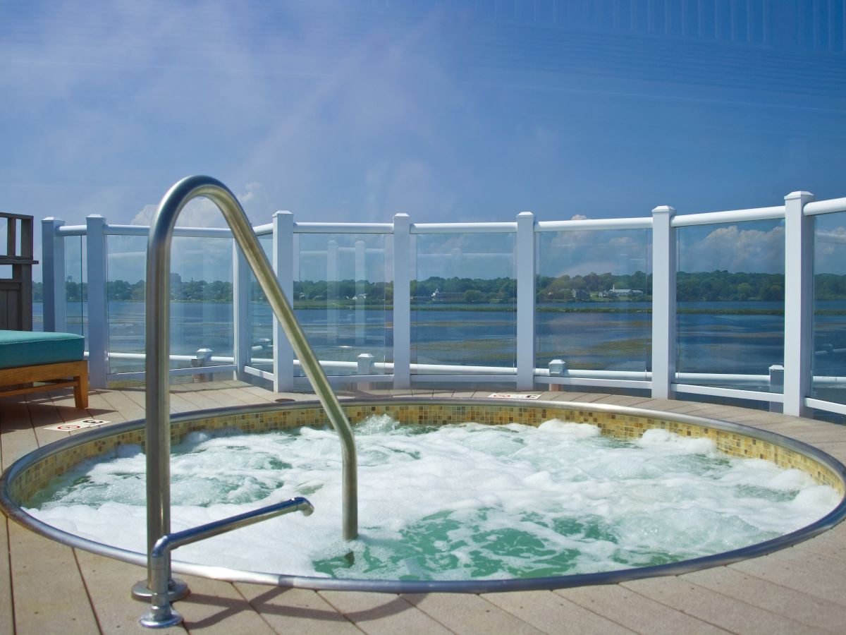 An outdoor hot tub on a deck with a metal handrail, glass railing, and a lake or ocean beyond it, under a clear blue sky.