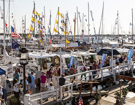 A busy marina packed with sailboats and flags, with people walking along the dock in bright daylight.