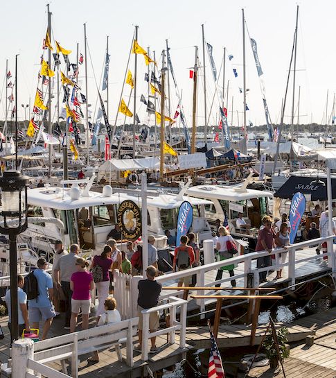 A busy marina packed with sailboats and flags, with people walking along the dock in bright daylight.