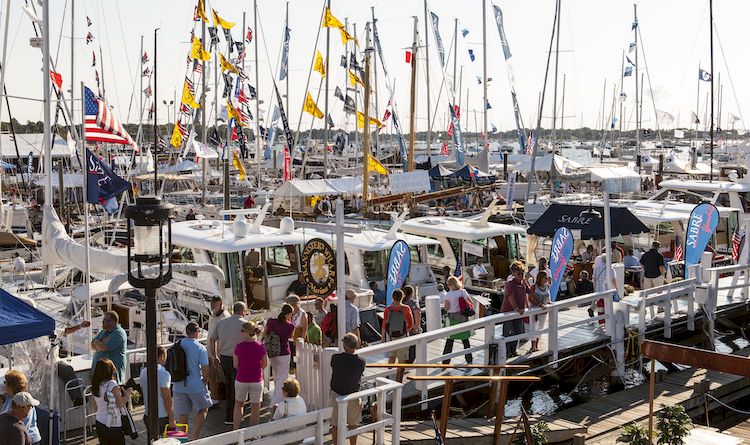 A busy marina packed with sailboats and flags, with people walking along the dock in bright daylight.