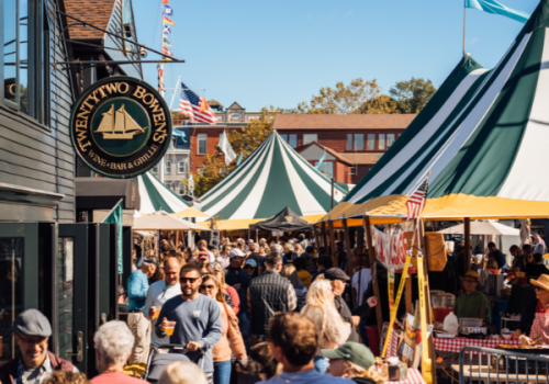 Crowds mill at an outdoor market with tents and flags near a bar-and-grill storefront.