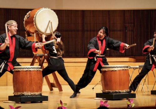 A group of performers in black and red outfits play taiko drums on a concert stage, with large drums and energetic poses.