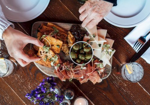 A group assembles a shared board with bread, prosciutto, olives, cheese, and microgreens on a wooden table, ready to eat.