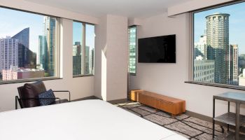 A modern hotel room with large windows offering a city view, a wall-mounted TV, a chair, and a wooden bench below the TV.