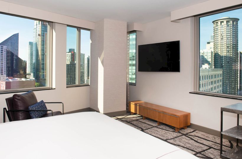 A modern hotel room with large windows offering a city view, a wall-mounted TV, a chair, and a wooden bench below the TV.