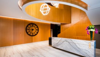 A modern lobby with a wooden spiral staircase, large clock on the wall, marble reception desk, and floral arrangement on the counter.