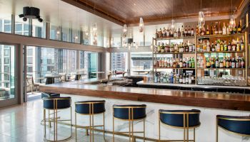 A modern bar with a wooden ceiling, high stools, and a wide selection of bottles on shelves. Large windows offer city views in the background.