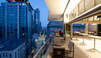 A modern balcony at night features seating and tables, overlooking a city street with tall buildings. Evening lights create a vibrant atmosphere.