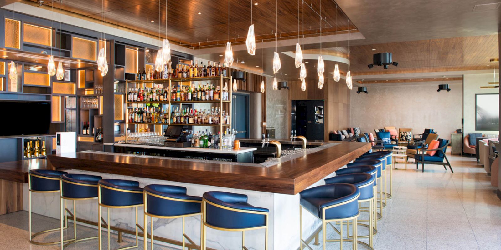 The image shows a modern bar with a well-stocked liquor shelf, stylish bar stools, warm lighting, and a spacious, contemporary seating area.