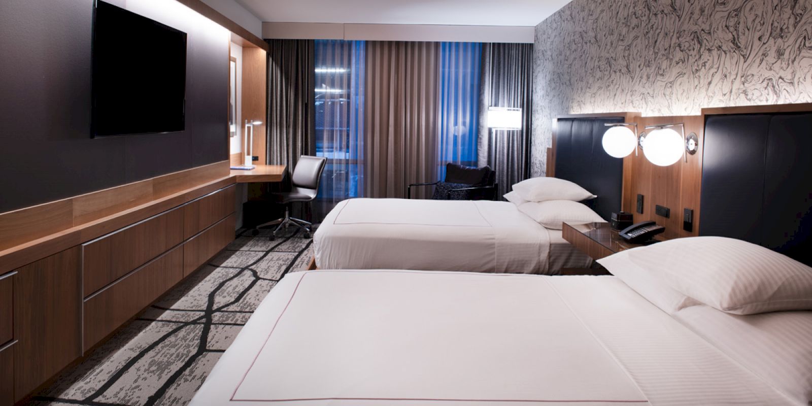 A modern hotel room with two beds, a desk, chair, and TV on the wall, featuring stylish decor and large windows with curtains.