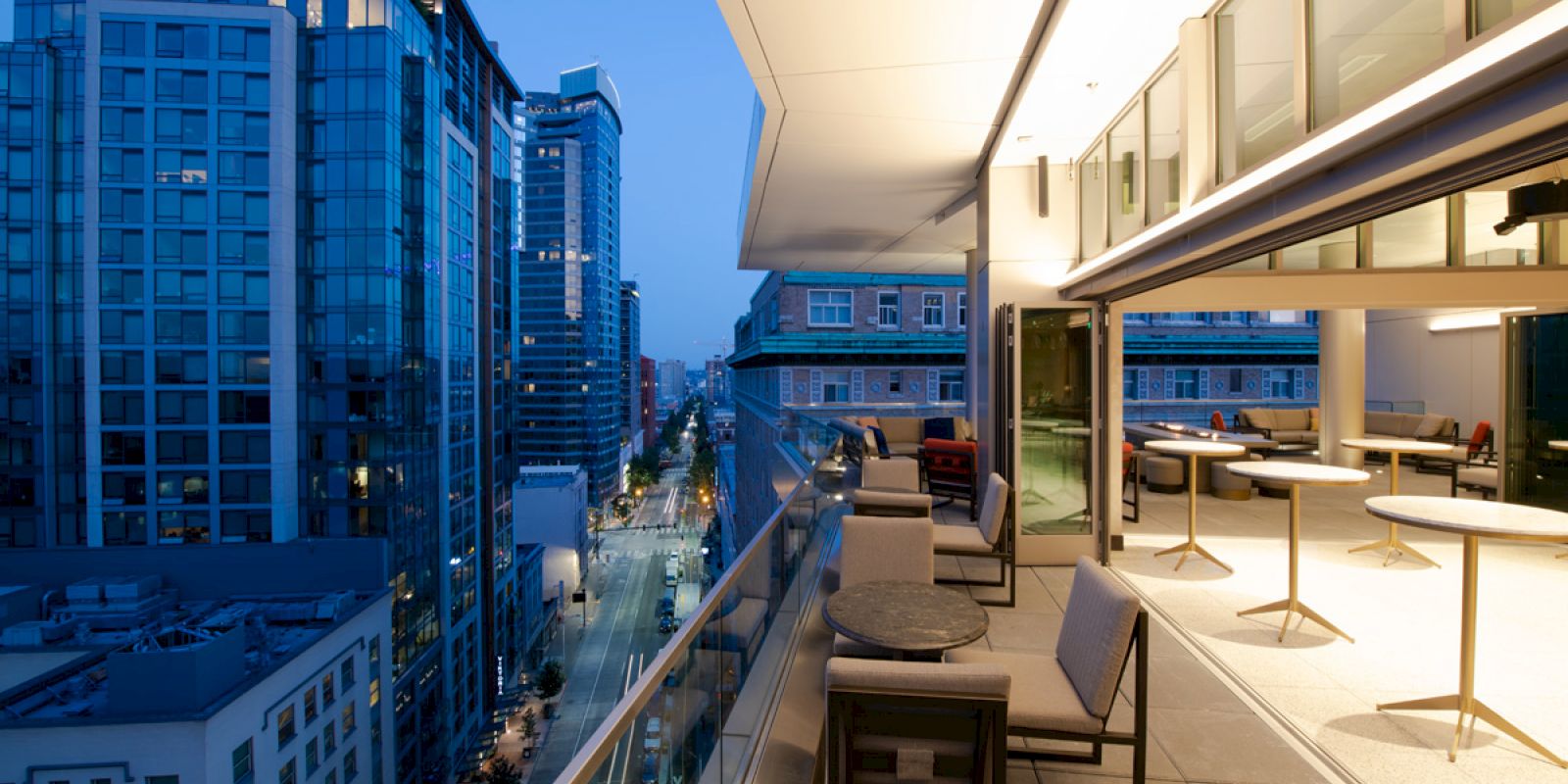 A modern, open-air balcony with cushioned chairs and tables overlooks a cityscape with lit buildings at dusk.