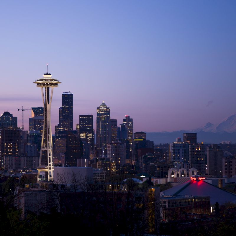 A city skyline at dusk featuring a prominent tower, with mountains visible in the background and illuminated buildings in the foreground.